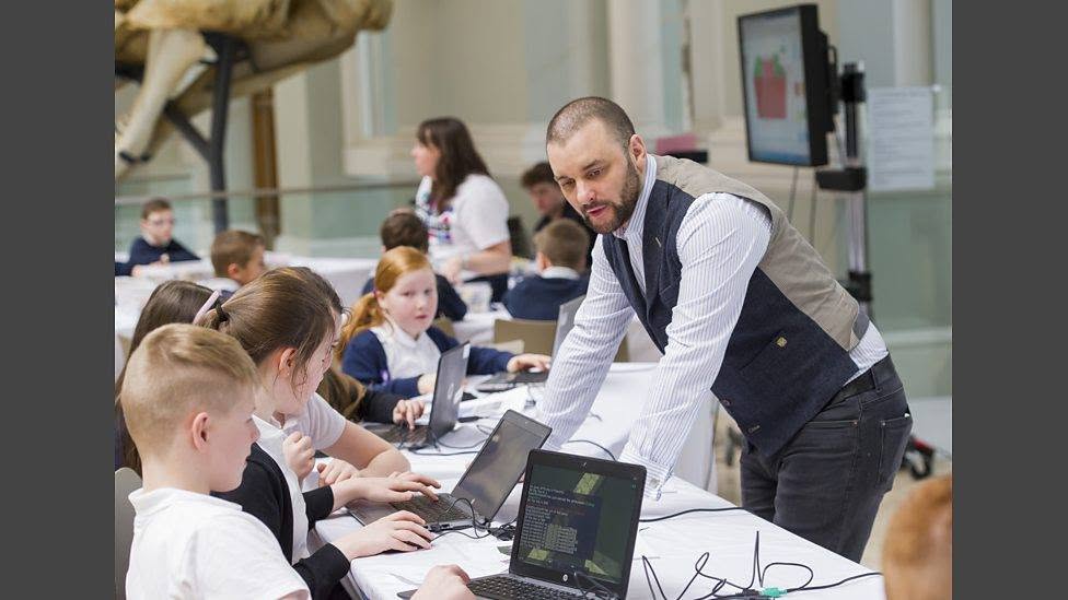 A facilitator leading a coding workshop with students at laptops in a museum setting.